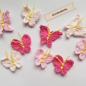 May include: A collection of handmade crochet butterflies in shades of pink and white, with yellow antennae. The butterflies are arranged on a white surface. The text "KristineKeita" is on a small tag.