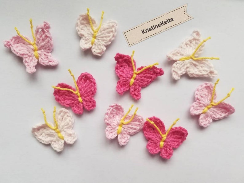 May include: A collection of handmade crochet butterflies in shades of pink and white, with yellow antennae. The butterflies are arranged on a white surface. The text "KristineKeita" is on a small tag.