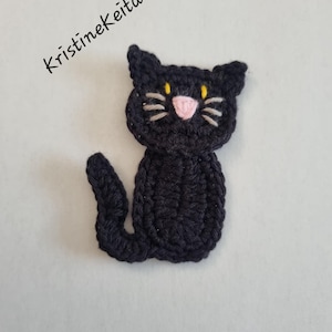 May include: A black crocheted cat applique featuring yellow embroidered eyes, a pink stitched nose, and grey whiskers. The text Kristinekeita is printed in black in the upper left corner of the image, positioned above the cat figure.