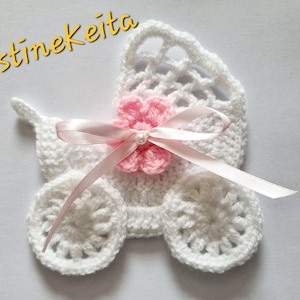 Può includere: Un passeggino bianco all'uncinetto con un fiore rosa e un nastro rosa. Il passeggino è un artigianato fatto a mano.