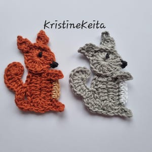 Peut inclure: Deux appliqués en forme d'écureuil crochetés. L'un est orange avec un ventre beige, l'autre est gris avec un ventre blanc. Les deux ont des yeux et un nez noirs. Le texte "Kristinekeita" est au-dessus des écureuils.