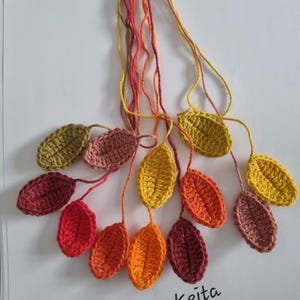 May include: A collection of twelve crocheted leaves in various autumn colours including yellow, orange, red, and brown, each attached to a matching yarn string. The leaves are arranged in a cluster, and the text Kristinekeita is printed below.