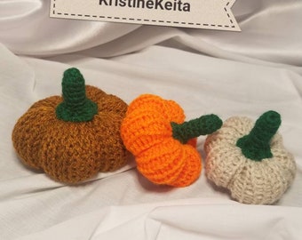 May include: Three crocheted pumpkins in different colors: brown, orange, and white. The pumpkins have green stems and are arranged on a white surface.