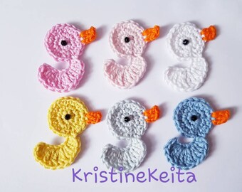 May include: Six handmade crochet duck appliques in various colors: pink, light pink, white, yellow, gray, and blue. Each duck has an orange beak and a black eye. The appliques are arranged on a white surface.