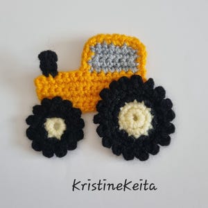 May include: A yellow, grey, and black crochet tractor applique with a small black chimney, a grey window section, and two large black wheels with cream-coloured centres. The text KristineKeita is printed in black below the tractor.