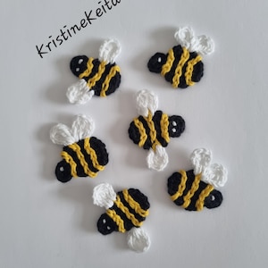 May include: Six small crocheted bee appliques featuring black and yellow striped bodies with white wings. The text KristineKeita is printed in black in the upper left corner. Each bee is crafted with yarn and displays a consistent design.