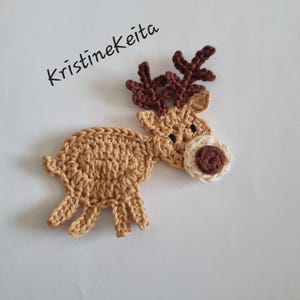 May include: A crocheted reindeer applique in shades of tan and brown, with a white and brown nose. The reindeer has detailed antlers and is set against a white background. The text "Kristinekeita" is written in black cursive.