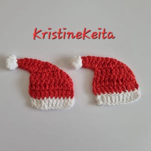 May include: Two handmade, red and white crocheted Santa hats with white pom-poms. The hats are on a white surface, with the name "Kristinekeita" in red script above them. These festive hats are perfect for holiday crafts.
