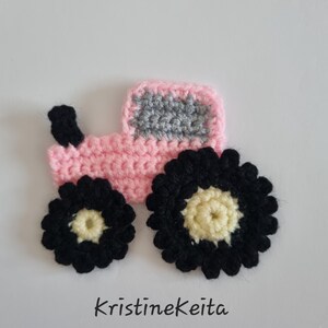 May include: A crochet tractor applique featuring a light pink body, a grey window section, and a black exhaust pipe. The tractor has two wheels with black outer edges and cream-colored centers. The text KristineKeita is printed below the tractor.