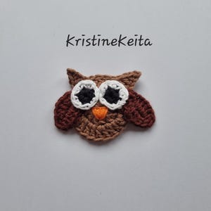 May include: A crocheted owl applique in shades of brown, with white and black eyes, and an orange beak. The owl has pointed ear tufts and wings. The text "kristinekeita" is above the owl.