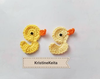 May include: Two crocheted yellow duck ornaments with orange beaks and black eyes. The ducks are on a white background with a brown banner that says "KristineKeita".