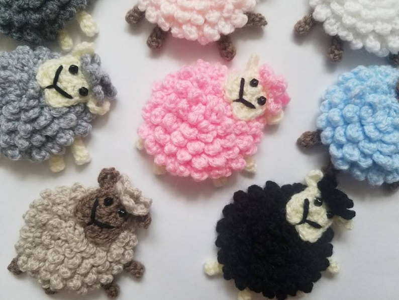 May include: A collection of crocheted sheep in various colors, including pink, blue, gray, white, and black. Each sheep has a fluffy, textured body and a cute face with black eyes and a smile.