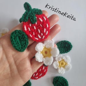 May include: Crocheted strawberry and flower appliques in red, white, yellow, and green yarn. The strawberry features white embroidered seeds and a green stem. The flowers have white petals and yellow centres. The text Kristinekeita is visible.