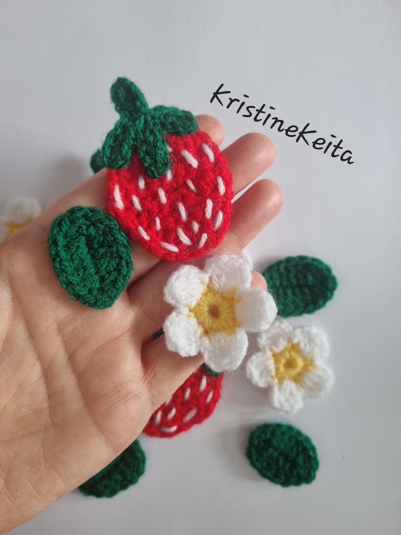 May include: Crocheted strawberry and flower appliques in red, white, yellow, and green yarn. The strawberry features white embroidered seeds and a green stem. The flowers have white petals and yellow centres. The text Kristinekeita is visible.