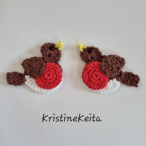 May include: Two crocheted robin bird appliques featuring brown heads and wings, bright red chests, white underbellies, and small yellow beaks. Each bird has a black bead eye. The text KristineKeita is printed in black below the two birds.