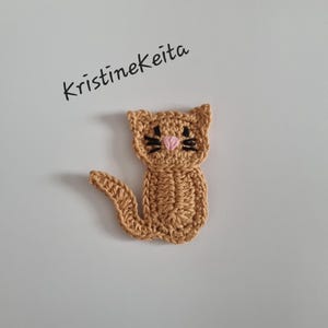 May include: A crocheted cat applique in shades of tan, with black whiskers and a pink nose. The cat has a curved tail and is set against a white background. The text "kristinekeita" is written in black cursive.