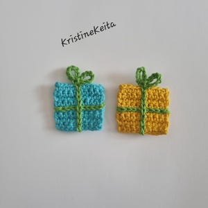 Pode incluir: Dois ornamentos quadrados de presente feitos em croch&ecirc;, um azul claro e outro amarelo, cada um com uma fita e um la&ccedil;o verdes feitos em croch&ecirc; no topo. O texto KristineKeita est&aacute; impresso em preto acima. Itens feitos de fio texturizado.