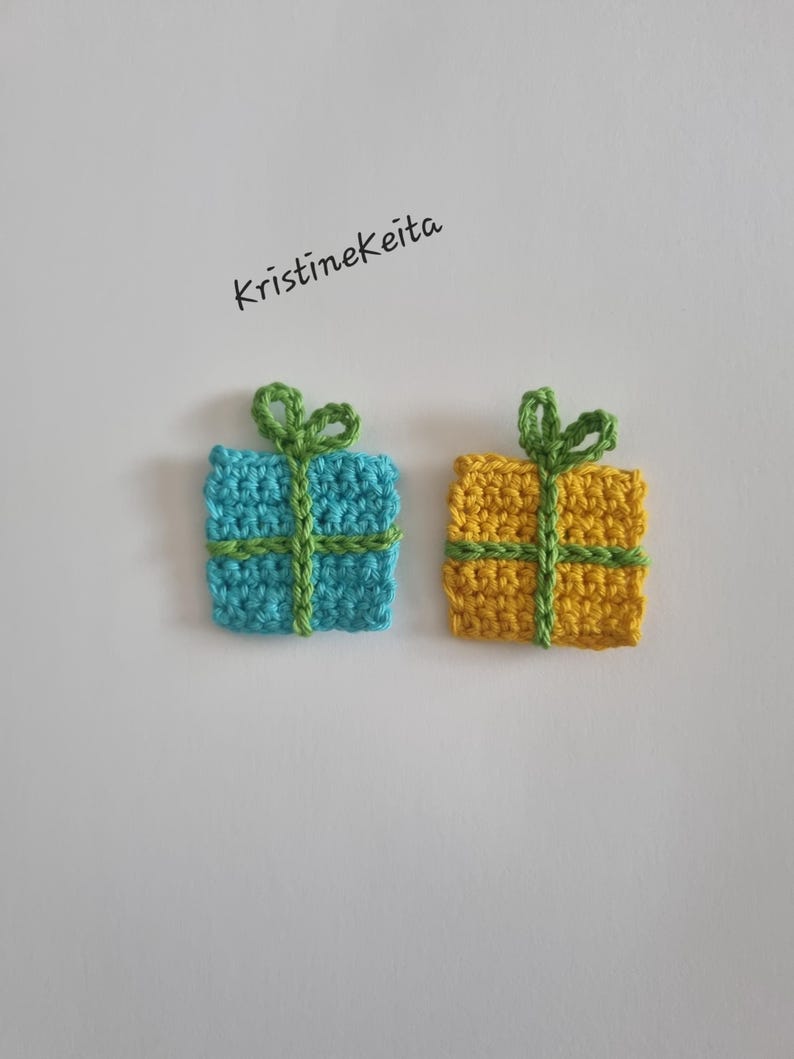 Pode incluir: Dois ornamentos quadrados de presente feitos em croch&ecirc;, um azul claro e outro amarelo, cada um com uma fita e um la&ccedil;o verdes feitos em croch&ecirc; no topo. O texto KristineKeita est&aacute; impresso em preto acima. Itens feitos de fio texturizado.