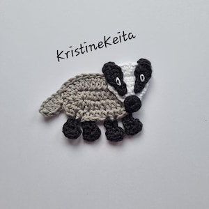 May include: A handmade crochet badger in grey, black, and white. The badger features a grey body, black feet, and a black and white face. The text "KristineKeita" is written in black cursive above the badger.