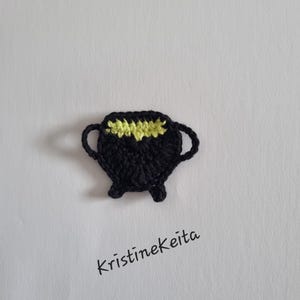 May include: A black crocheted cauldron appliqué with two small handles and three short legs. The top of the cauldron features a bright yellow crocheted accent representing liquid. The text Kristinekeita is printed in black below the object.