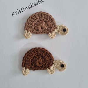 Könnte beinhalten: Zwei gehäkelte Schildkröten-Applikationen mit dem sichtbaren Text Kristinekeita. Die obere Schildkröte hat einen hellbraunen Panzer, die untere einen dunkelbraunen. Beide haben cremefarbene Köpfe mit schwarzen Augen und cremefarbene Gliedmaßen.