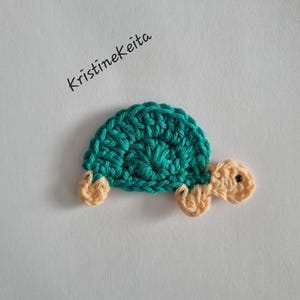 May include: A small, flat, crocheted turtle applique featuring a teal shell and light peach-coloured head and limbs. The turtle has a single black bead for an eye. The text KristineKeita is printed in black ink above the crocheted turtle figure.