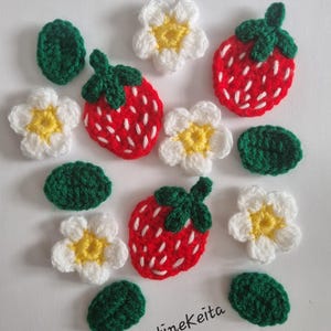 May include: A collection of crocheted appliques featuring three red strawberries with green tops and white seed details, four white flowers with yellow centres, and four solid green leaves. The text Kristinekeita is printed in black.