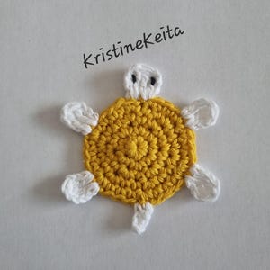Op de afbeelding: Een kleine, platte gehaakte schildpadapplicatie met een rond geel schild, een witte kop met twee zwarte geborduurde ogen, vier witte flippers en een kleine witte staart. De tekst KristineKeita staat in het zwart boven de schildpad.