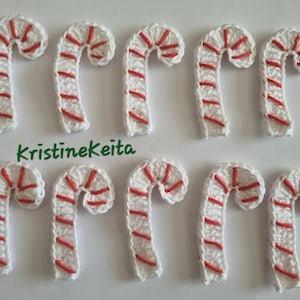 Könnte beinhalten: Zehn gehäkelte Zuckerstangen. Jede Zuckerstange ist weiß mit roten Streifen und einer gebogenen Form. Der Text "kristinekeita" steht in grüner Schrift unter den Zuckerstangen.