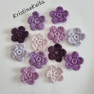 May include: A selection of crocheted flower appliques in shades of purple, lavender, and grey. The handmade flowers are displayed on a white surface. The text "Kristinekeita" is in the upper left corner.