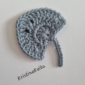 May include: A light gray crochet applique shaped like a stingray with a rounded body, a long tail, and two small black circular eyes. The text Kristinekeita is printed in black in the lower left corner of the image.