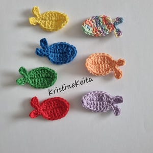 Könnte beinhalten: Sieben gehäkelte Fische in verschiedenen Farben: Gelb, Blau, Grün, Rot, Orange, Lila und ein mehrfarbiger Fisch. Jeder Fisch hat einen runden Körper und einen kleinen Schwanz. Der Text "Kristinekeita" ist sichtbar.