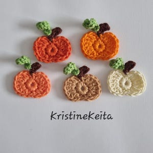May include: Five small, flat, crocheted pumpkin appliques arranged in a group. Each pumpkin features a round, textured body in shades of orange, tan, or cream, with a small brown stem and a light green leaf. The text KristineKeita is printed below.