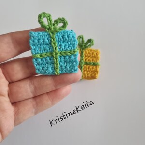 Pode incluir: Dois pequenos ornamentos quadrados de presente feitos em croch&ecirc;. Um &eacute; azul-claro com um la&ccedil;o de fio verde, e o outro &eacute; amarelo com um la&ccedil;o de fio verde. O texto Kristinekeita est&aacute; impresso em preto sobre o fundo branco.