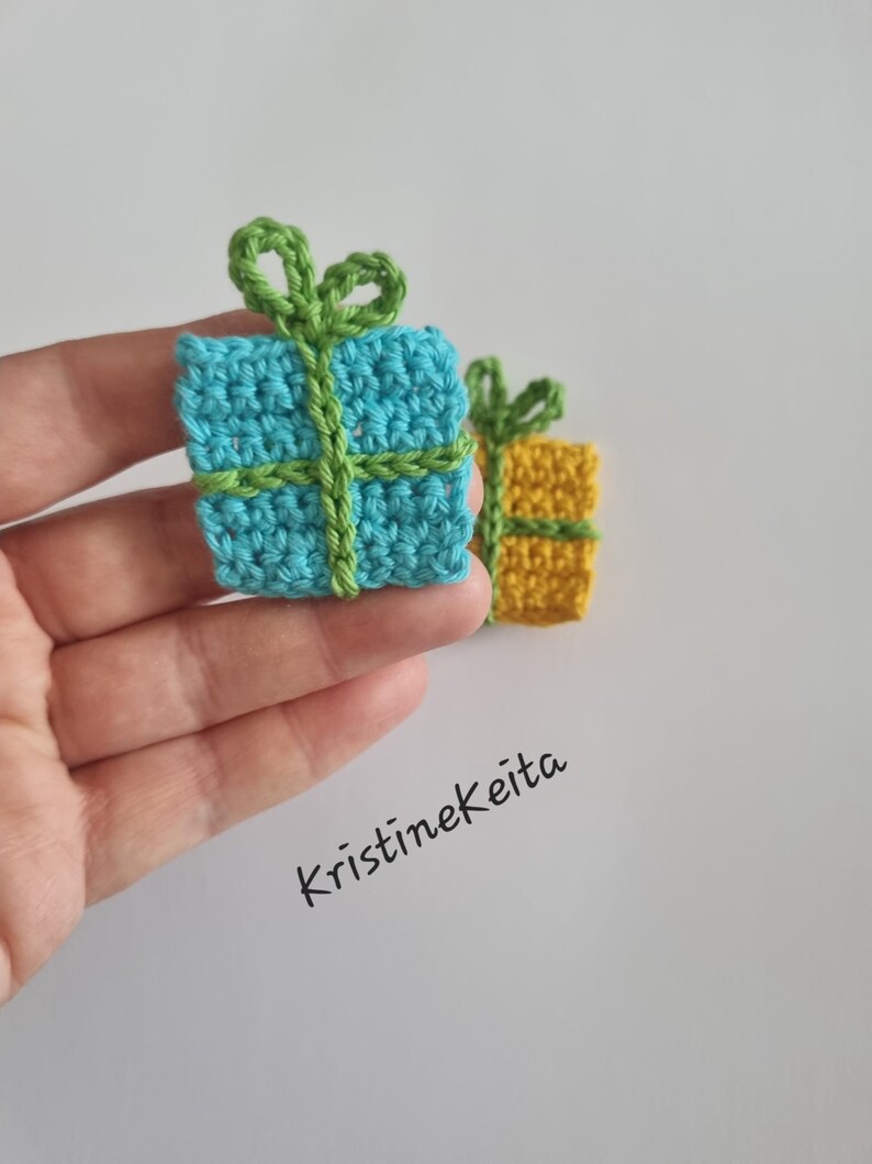 Pode incluir: Dois pequenos ornamentos quadrados de presente feitos em croch&ecirc;. Um &eacute; azul-claro com um la&ccedil;o de fio verde, e o outro &eacute; amarelo com um la&ccedil;o de fio verde. O texto Kristinekeita est&aacute; impresso em preto sobre o fundo branco.