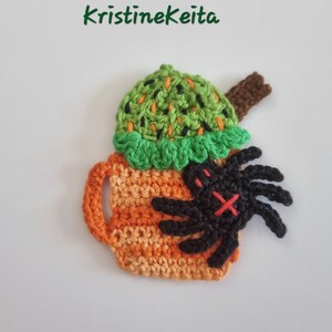May include: A crocheted Halloween-themed design of a pumpkin spice latte with a spider. The latte is orange and yellow, topped with green and black whipped cream. A black spider with a red 'X' is on the side. The text "Kristinekeita" is above the latte.