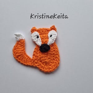 May include: A crocheted orange fox applique with white eye and tail tip details, and a black nose. The text "Kristinekeita" is above the fox. Ideal for adding to clothing or accessories, this is a handmade item.