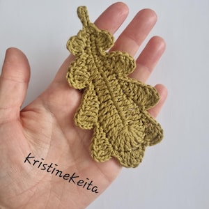 May include: A crocheted oak leaf appliqué in a muted olive green colour. The leaf features a detailed central vein and lobed edges, with a small loop at the top for hanging. The text Kristinekeita is printed in black on the lower left side.