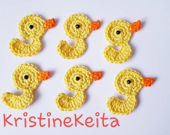 May include: Six yellow crocheted duck ornaments with orange beaks and black eyes. The ducks are arranged in two rows of three. The text "KristineKeita" is written in pink below the ducks.