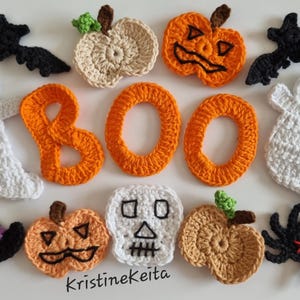 Op de afbeelding: Een verzameling gehaakte Halloween-decoraties waaronder twee witte spoken, twee zwarte vleermuizen, een zwarte spin, een zwarte heksenhoed met een gele gesp, een witte schedel en vier pompoenen in oranje en beige, met de oranje gehaakte letters B, O, O.