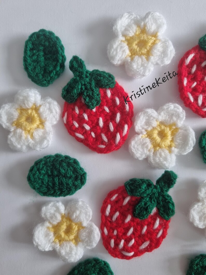 May include: A collection of crocheted appliques featuring red strawberries with white embroidered seeds and green leaves, white flowers with yellow centres, and solid green leaves. The text KristineKeita is visible in black script.