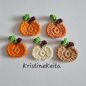 May include: Five small crocheted pumpkin appliques arranged in a group. Each pumpkin features a textured orange, cream, or tan body with a small green leaf and a brown stem. The text KristineKeita is printed in black below the crocheted items.