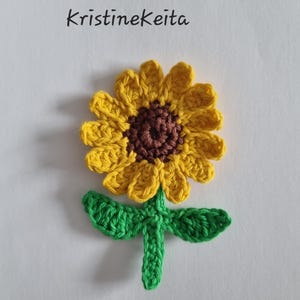 May include: A crocheted sunflower appliqué featuring a dark brown centre, bright yellow petals, and a green stem with two leaves. The text KristineKeita is printed in black above the flower. This textile piece is crafted with yarn.