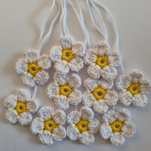 May include: A collection of nine small, handmade crochet flowers, each featuring five white petals and a bright yellow centre. Each flower is attached to a thin white string extending upwards. The text KristineKeita is printed at the bottom.