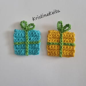 Pode incluir: Dois apliques de croch&ecirc; em formato de caixa de presente quadrada, um em azul brilhante e outro em amarelo, cada um com um detalhe de fita e la&ccedil;o de croch&ecirc; verde. O texto KristineKeita est&aacute; impresso em preto acima da caixa azul no fundo branco.