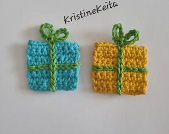 May include: Two square crochet gift box appliques, one in bright blue and one in yellow, each featuring a green crocheted ribbon and bow detail. The text KristineKeita is printed in black above the blue gift box on the plain white background.