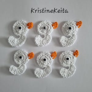 May include: Six white crocheted swan appliques arranged in two rows of three. Each swan features a small orange beak and a single black bead eye. The text KristineKeita is printed in black above the swan appliques on a plain white background.
