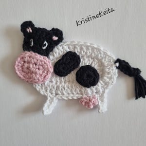 May include: A crochet cow appliqué featuring a white body with black spots, a black head, a pink snout, and a black tail. The text KristineKeita is printed in black above the cow. The design is crafted with visible yarn stitches in a flat, decorative style.