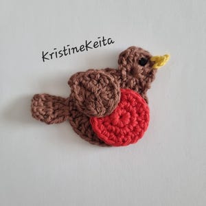 May include: A handmade crochet bird in shades of brown and red, with a yellow beak and a small black eye. The bird is set against a white background. The text "kristinekeita" is written in black cursive.
