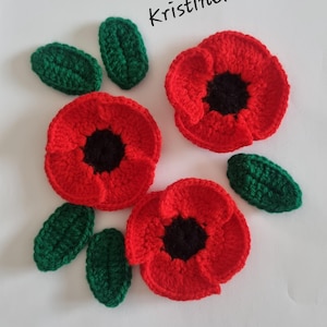 May include: Three crocheted red poppy flowers with black centres and four green crocheted leaves arranged on a white surface. The text Kristinekeita is printed in black in the upper right area of the composition, above the floral elements.